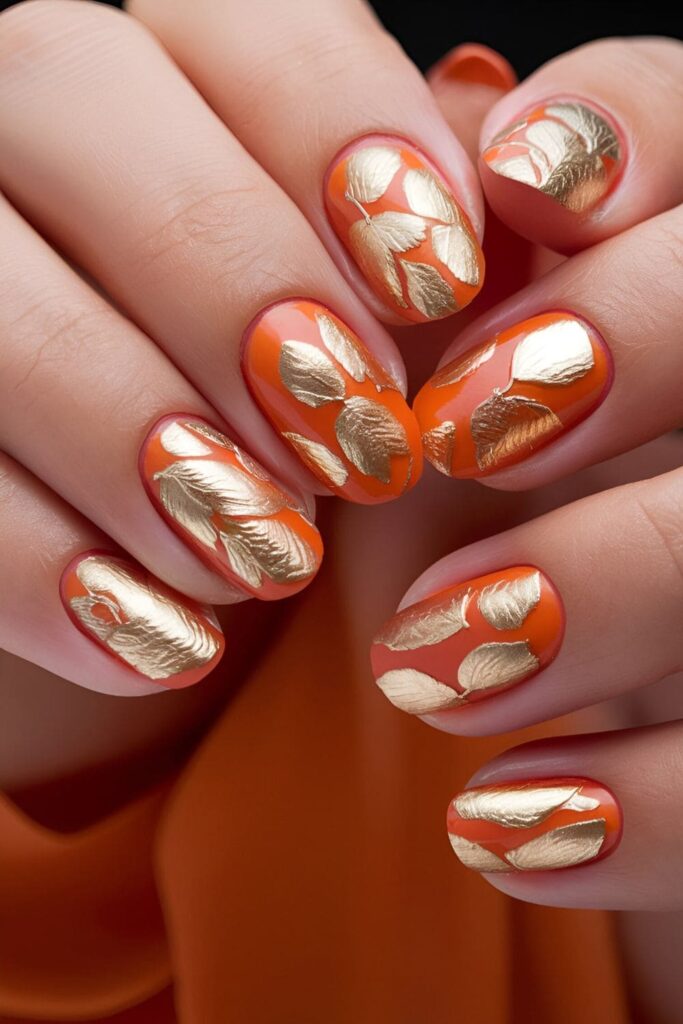 Elegant acrylic nails adorned with intricate gold leaf patterns on an orange base, luxurious and stylish nail art