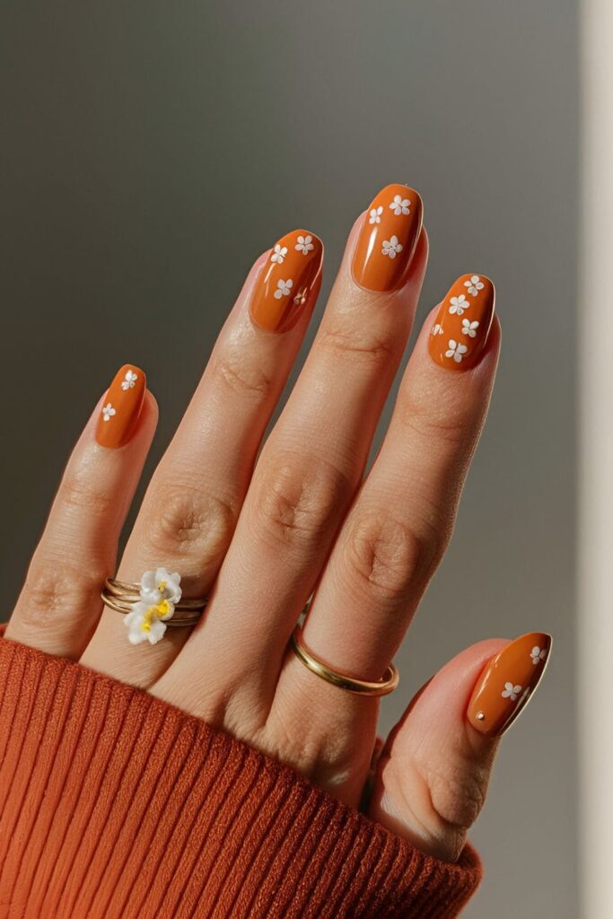 Soft, muted orange nails adorned with small white and yellow flowers, evoking fall foliage and blooms