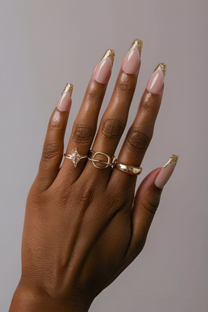 Dark-skinned hands with nude acrylic nails featuring gold foil accents and glitter tips