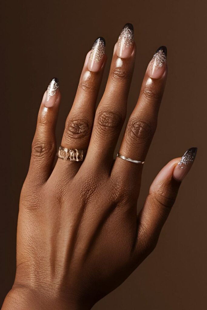 Dark-skinned hands with glitter gradient acrylic nails, featuring silver or gold glitter over a dark base