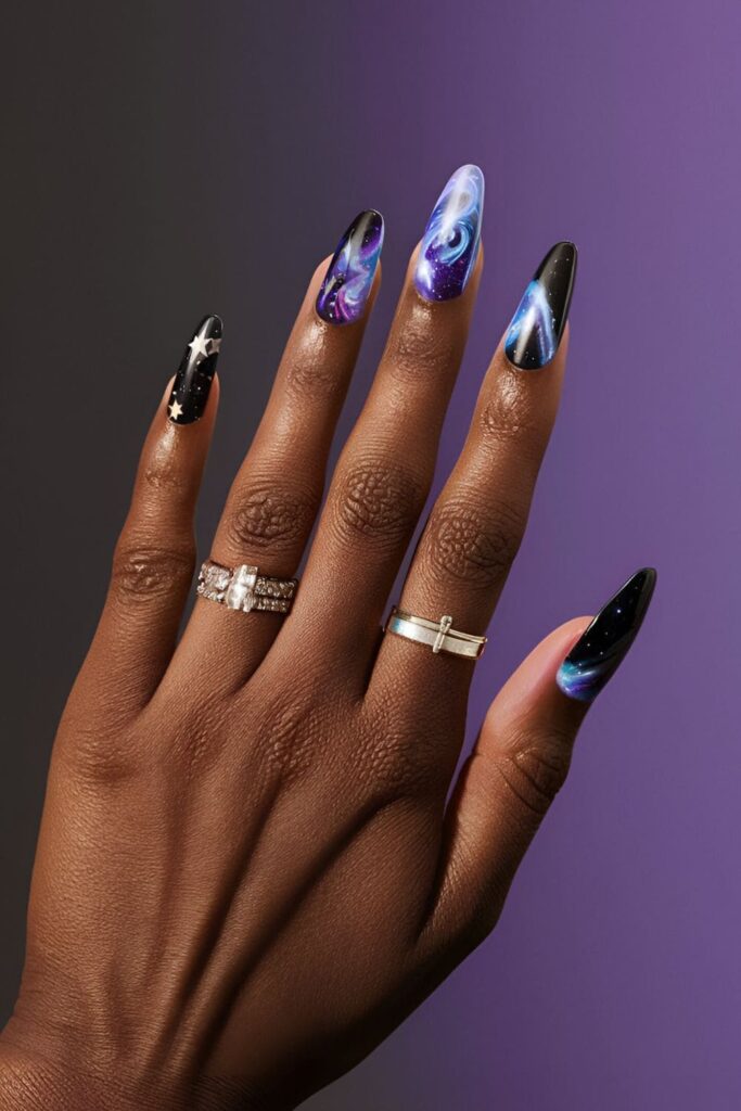 Dark-skinned hands with galaxy-themed acrylic nails, featuring swirls of blues, purples, blacks, and silver glitter stars