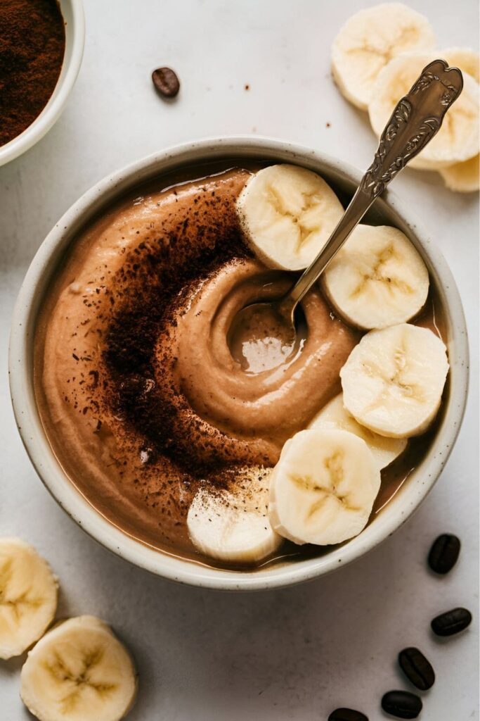 A smooth blend of mashed banana and coffee grounds in a bowl, with a spoon dipped in the mixture. Fresh banana slices and coffee beans are placed around the bowl