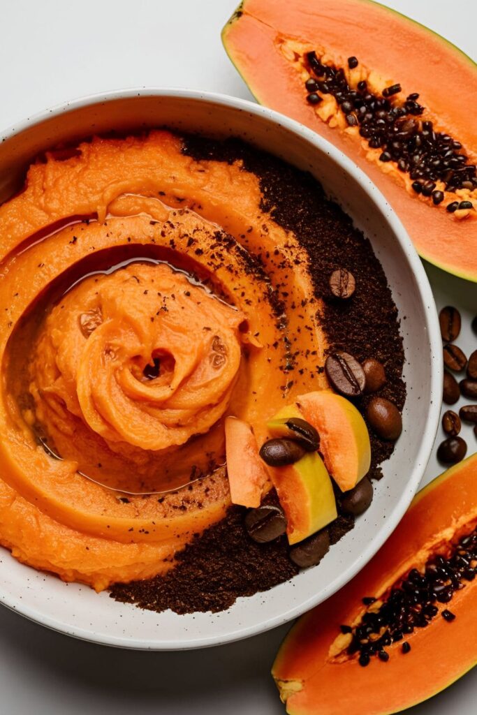 A vibrant orange mixture of mashed papaya and coffee grounds in a bowl, with papaya slices and coffee beans nearby. The texture is smooth with visible coffee granules