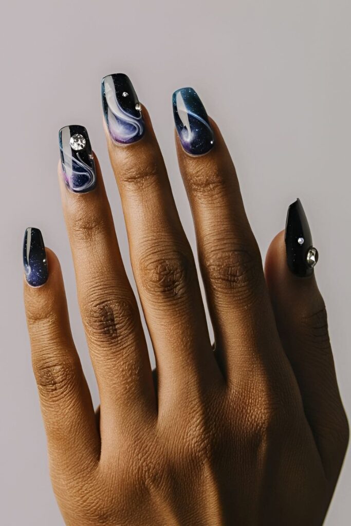 Galaxy-themed acrylic nails with dark blues, purples, and blacks, featuring swirls of white and rhinestones as stars for a mesmerizing celestial effect