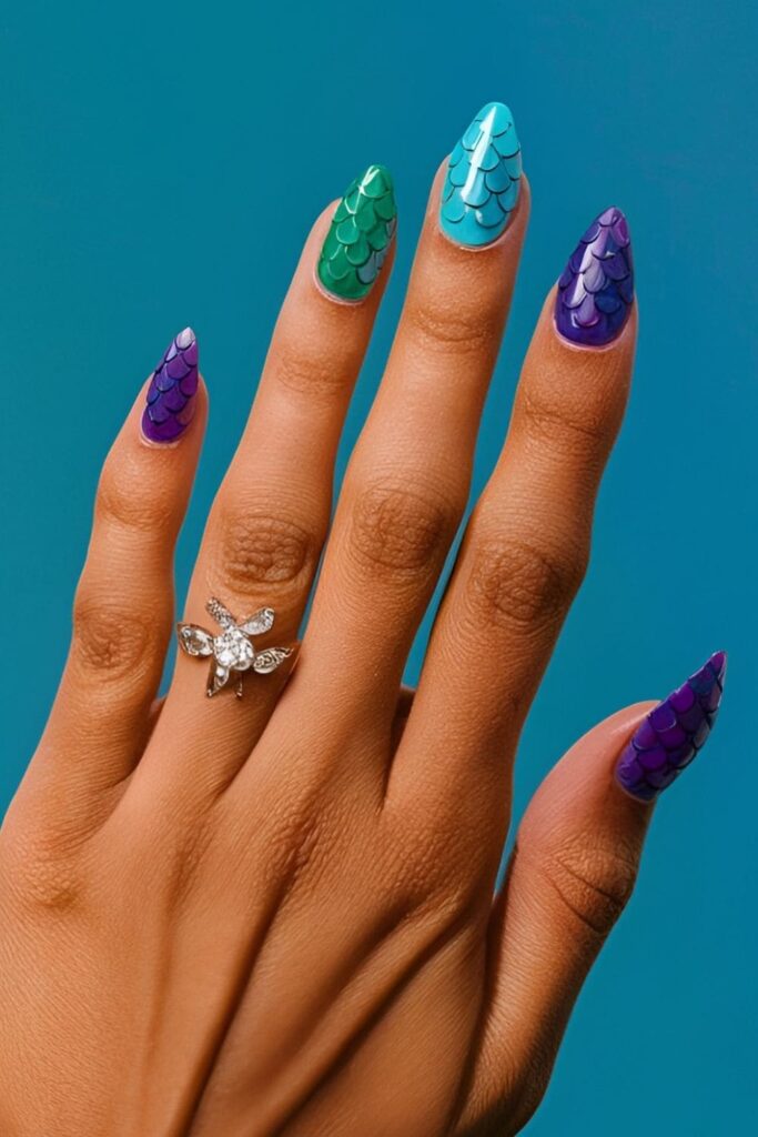 Mermaid-themed acrylic nails with shades of blue, green, and purple, featuring scale patterns and rhinestones representing underwater treasures