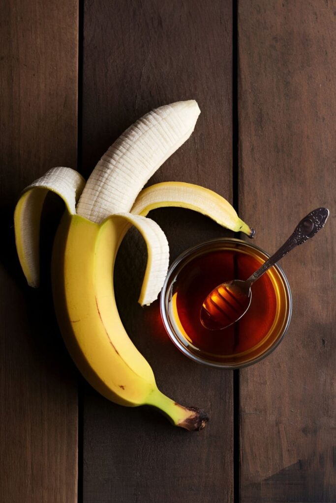 A ripe banana, peeled halfway, next to a small bowl of honey with a spoon on a rustic wooden table