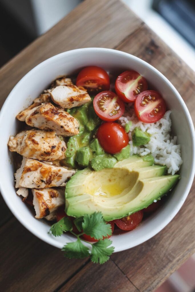 Grilled chicken and rice bowl topped with avocado, cherry tomatoes, and lime on a white plate