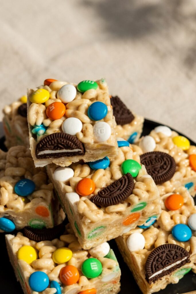 Rice Krispie treats mixed with M&Ms and Oreo chunks cut into squares and stacked on a tray